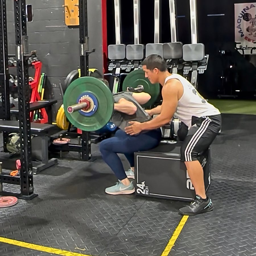 A woman squatting with a loaded barbell to a box while Coach Carlos spots her at Máquina Strength
