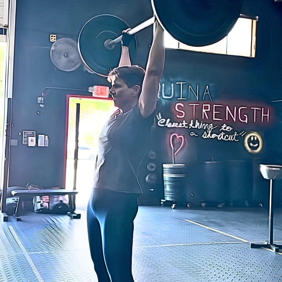 A woman in her sixties locking out a loaded barbell overhead at Máquina Strength in Palmetto Bay